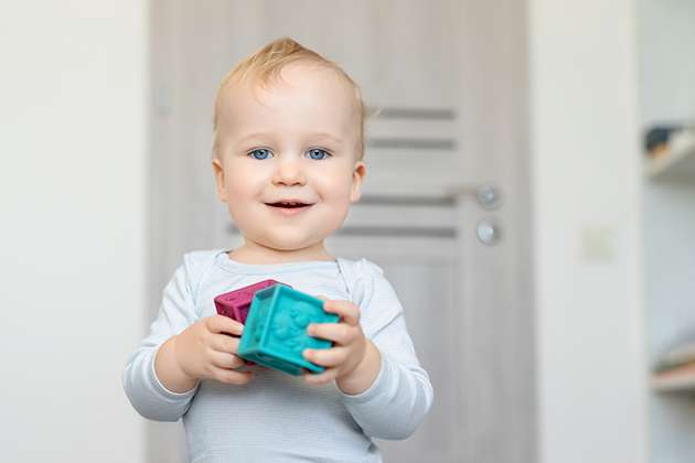 Un jeune enfant blond, souriant, tient deux cubes colorés en plastique (un turquoise et un violet) dans ses mains. Il est debout à l’intérieur, vêtu d’un body gris clair, et regarde vers l’objectif avec curiosité.