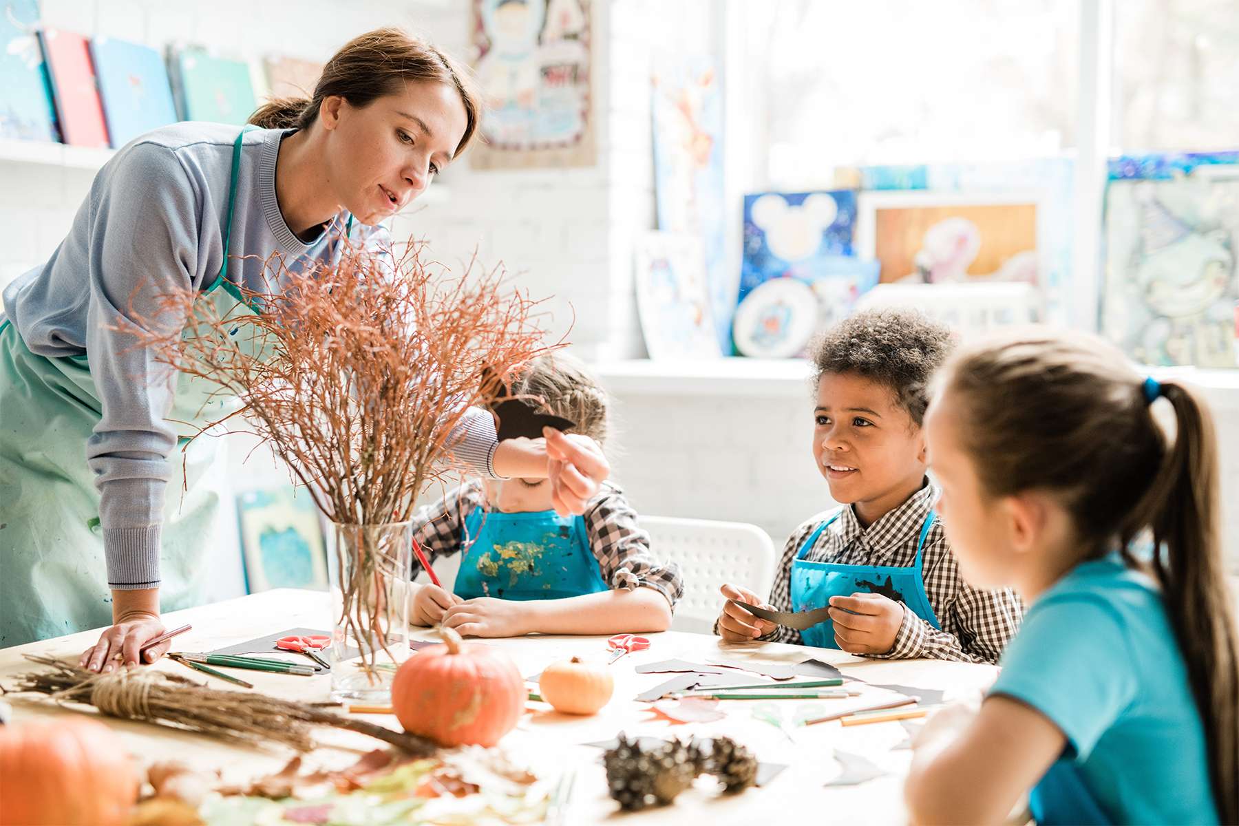 Une dame montre une chauve-souris en papier à trois enfants qui participent à un atelier artistique. Ils sont assis autour d'une table remplie de fournitures d'art et d’éléments naturels comme des branches, des citrouilles et des pommes de pin. Tous portent des tabliers de peinture et semblent concentrés et heureux.