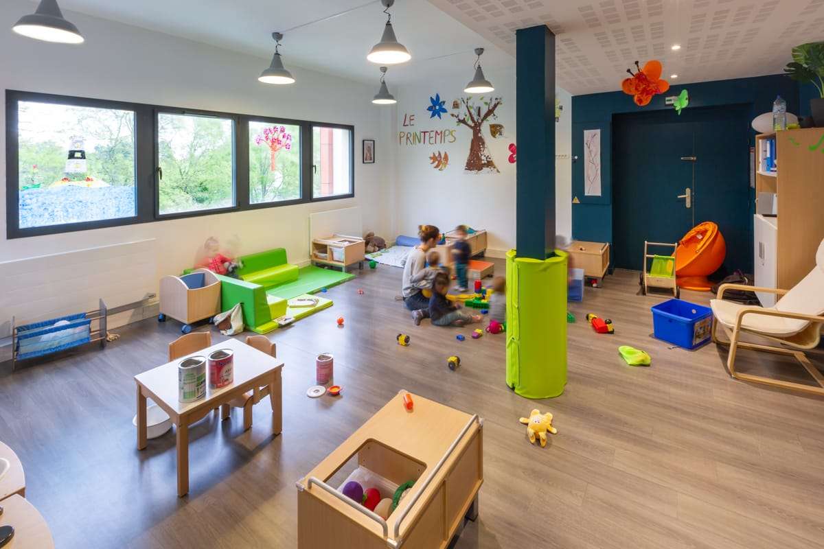Salle de jeux en crèche Une éducatrice joue avec plusieurs jeunes enfants dans une salle de crèche colorée, remplie de jouets, tapis et petits meubles adaptés.