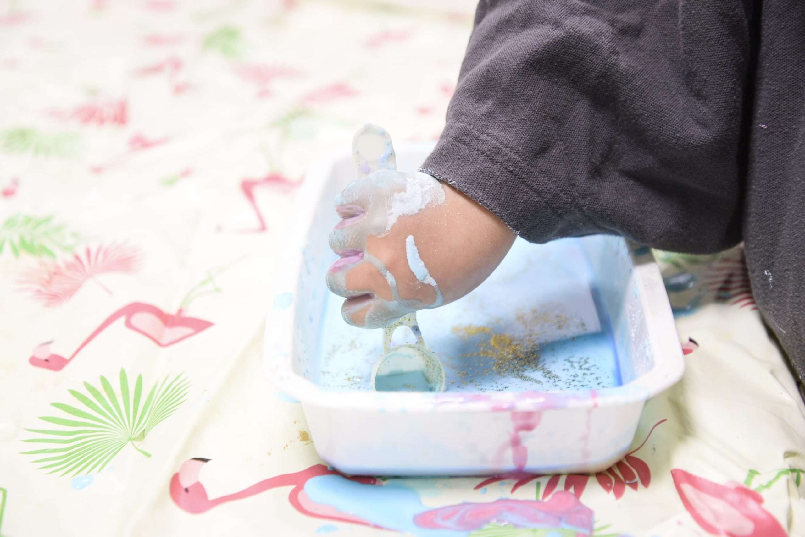 Petites mains créatives Gros plan sur la main d'un jeune enfant vêtue d'un pull gris, barbotant joyeusement dans un plateau rectangulaire rempli de peinture bleue et de paillettes dorées.