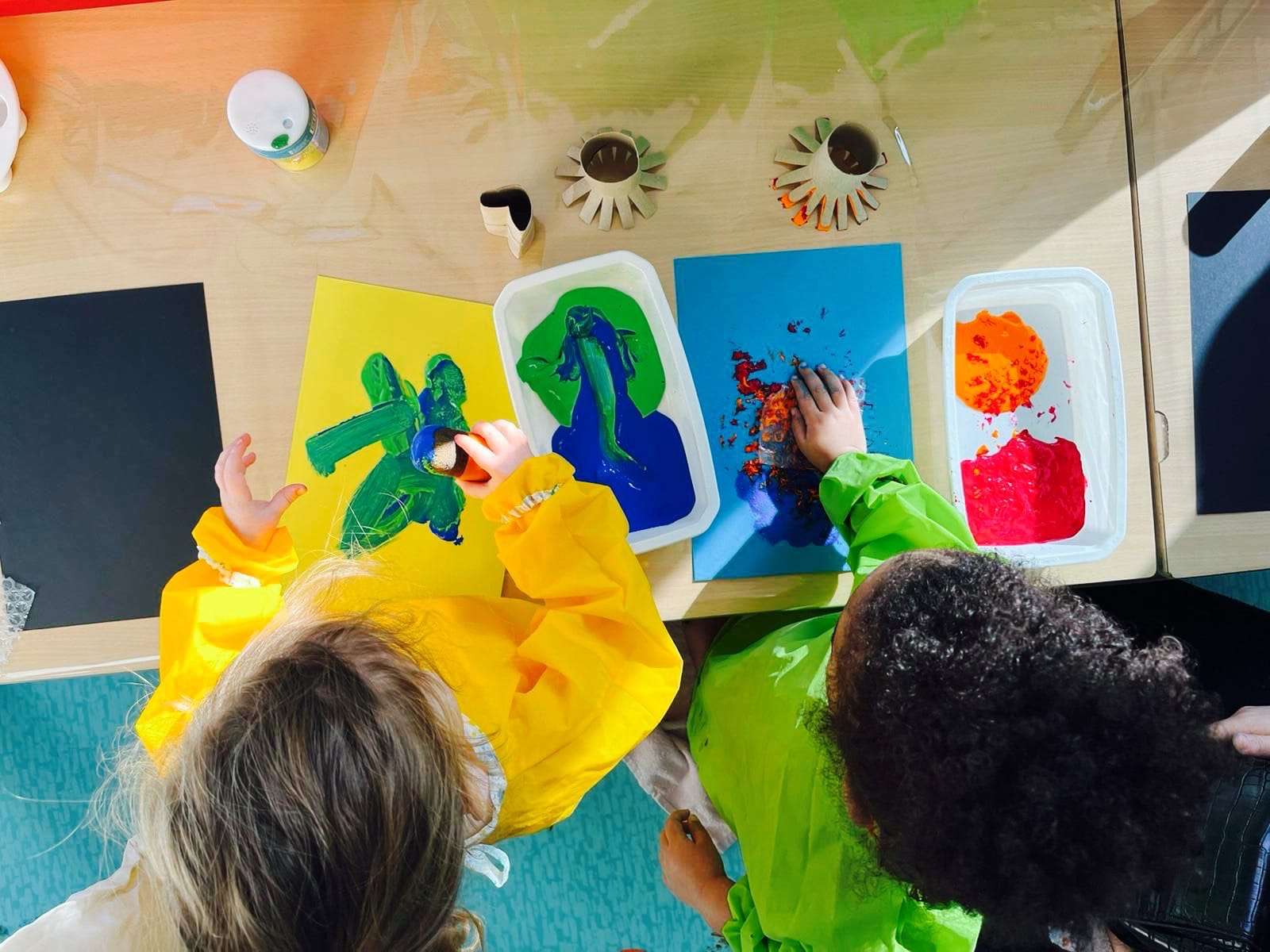 Vue d'en haut de deux enfants en bas âge, vus de dos, engagés dans une activité de peinture à une table en bois.