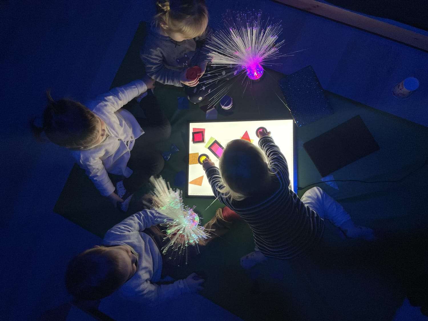 Quatre jeunes enfants explorent une table lumineuse et des jeux sensoriels colorés dans une salle Snoezelen à l’ambiance tamisée.