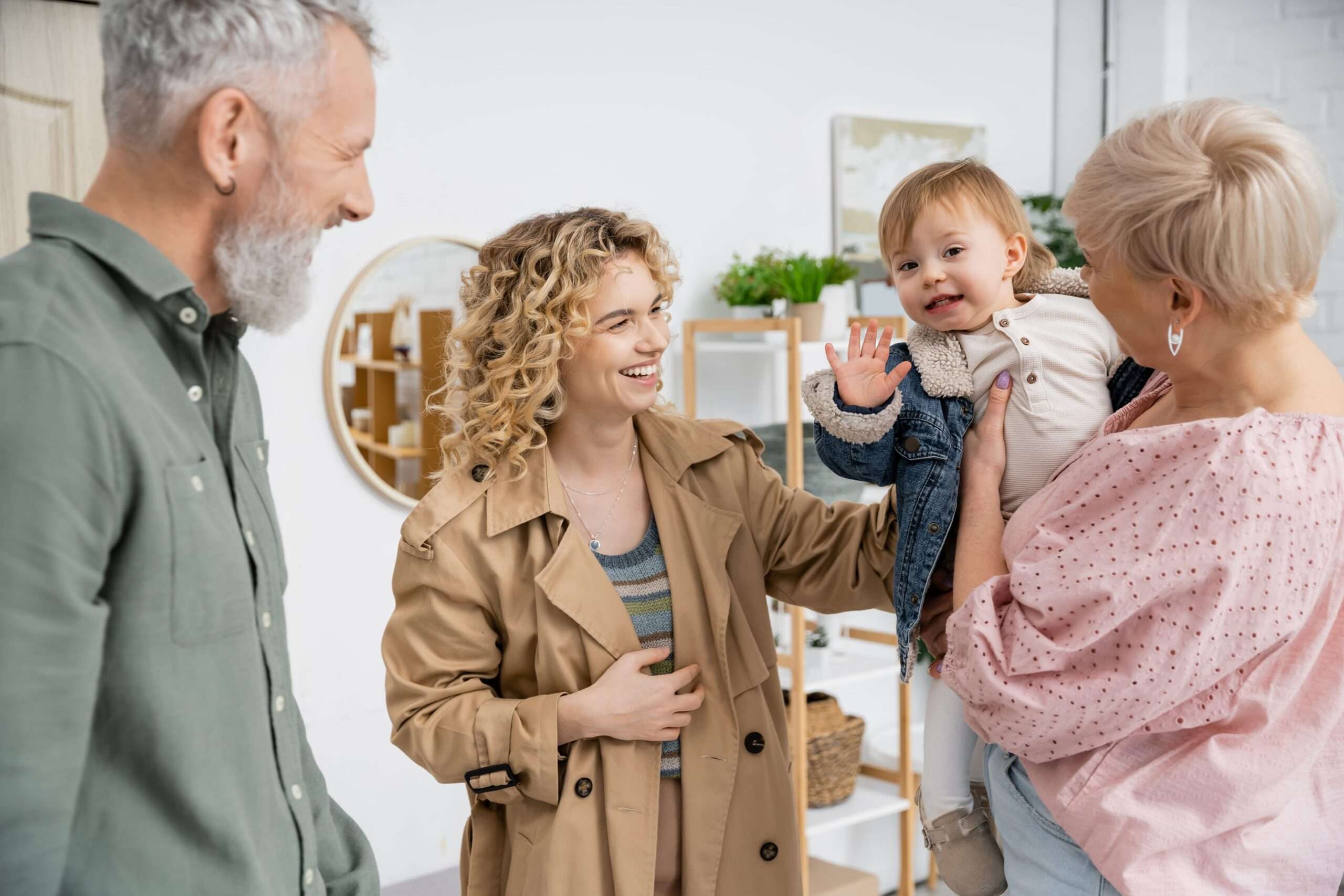 Une communication essentielle entre la famille et la crèche Un couple souriants interagit avec une jeune femme blonde aux cheveux bouclés, qui tient un enfant. Le bébé, vêtu d'une veste en jean, agite la main et sourit à la caméra.