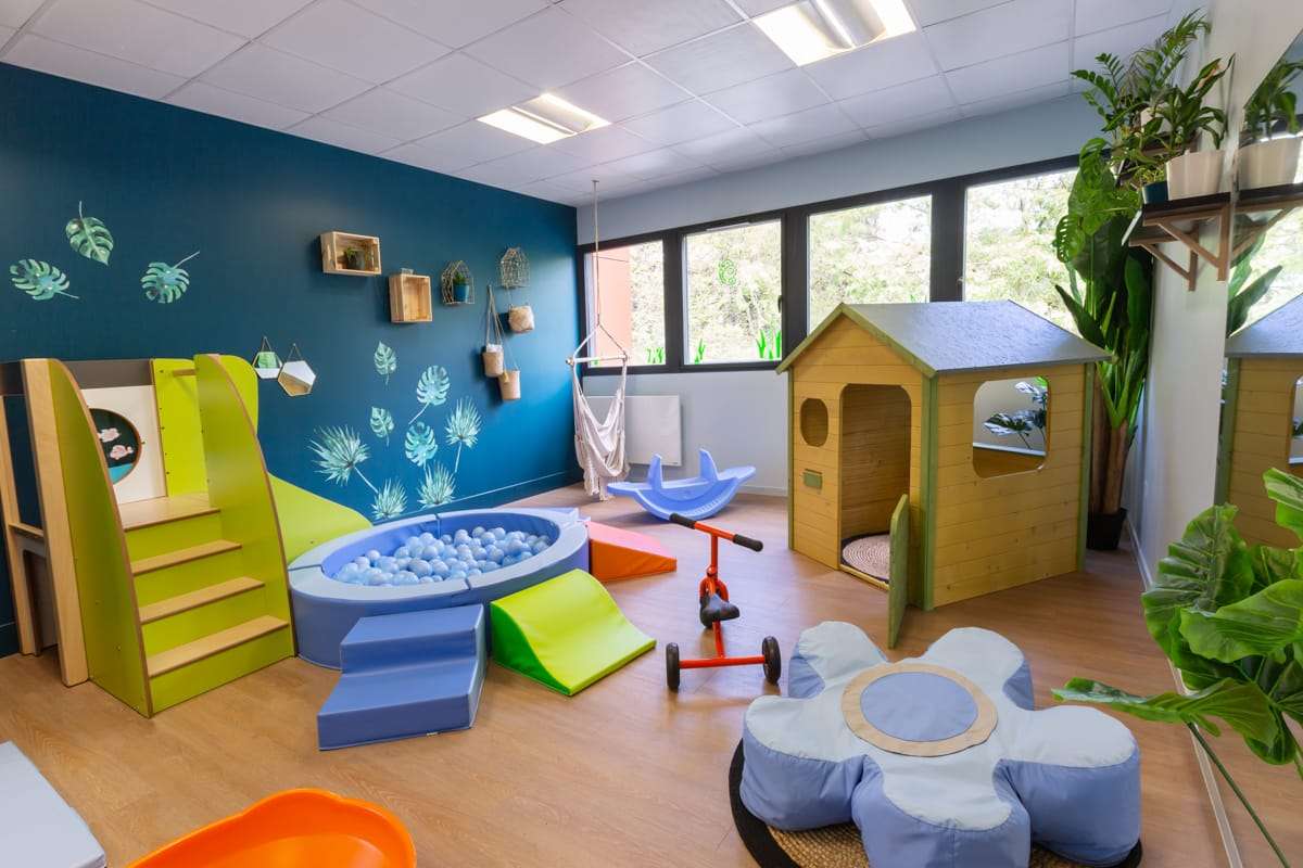 Vue intérieure d'une salle de motricité et de jeux dans la crèche de Saint-Martin. L'espace est très coloré et moderne, avec un mur peint en bleu sarcelle profond décoré de motifs végétaux et de petits étagères en bois.
