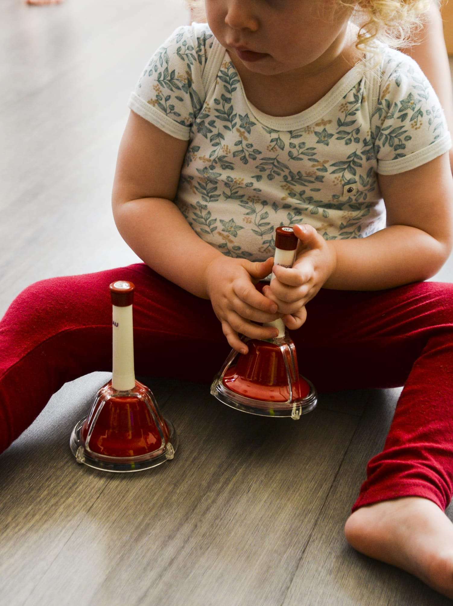 La découverte des sons Un enfant assis au sol tient deux petites cloches musicales rouges, découvrant les sons à la crèche.