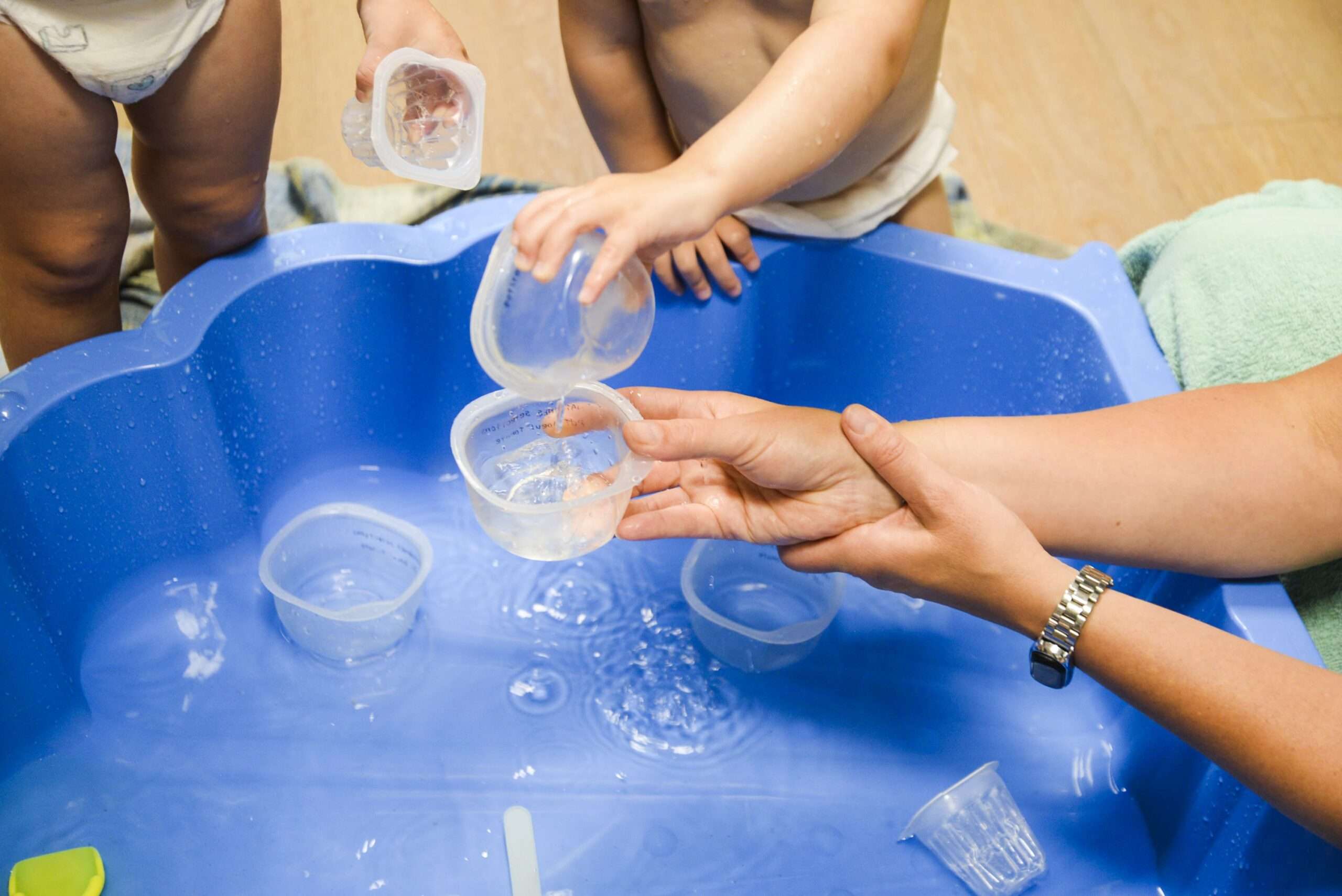 Des enfants en bas âge jouent avec de l’eau et des gobelets dans un bac bleu, sous la guidance d’un adulte.