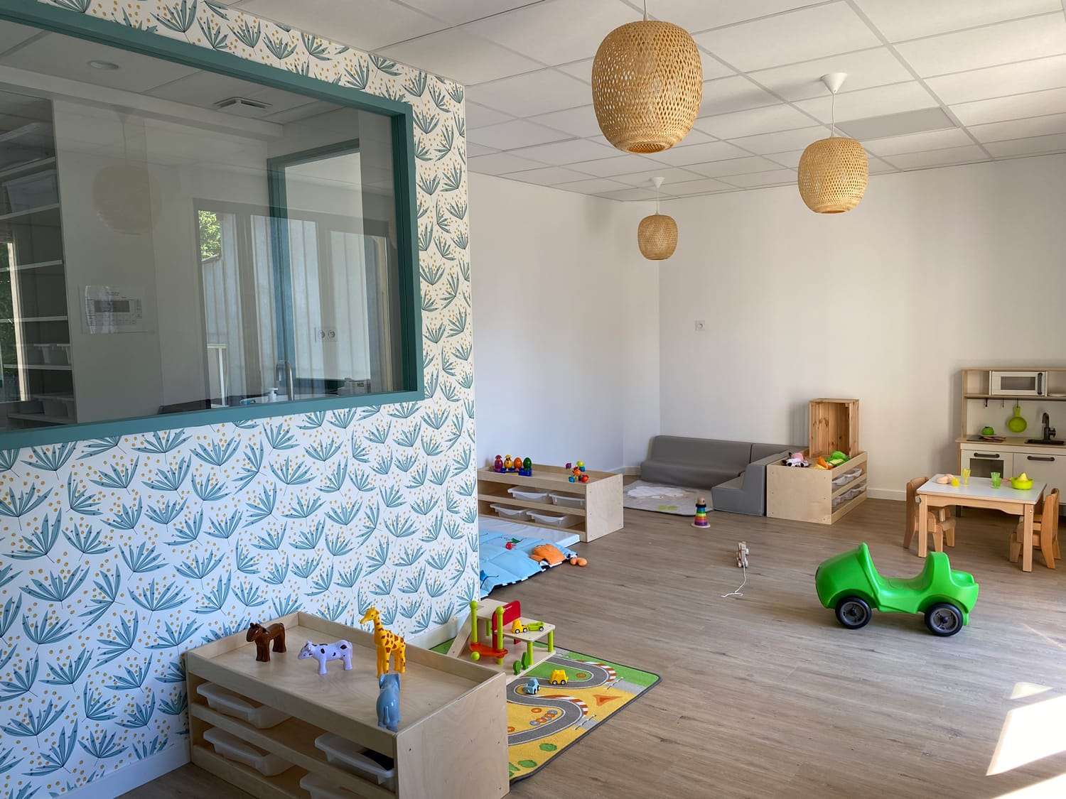 Vue panoramique d'une salle de jeux spacieuse et lumineuse dans une crèche. L'espace est aménagé avec des meubles bas en bois naturel pour le rangement des jouets, des tapis d'éveil au sol, un petit canapé gris et une dinette avec une table et des chaises pour enfants. 