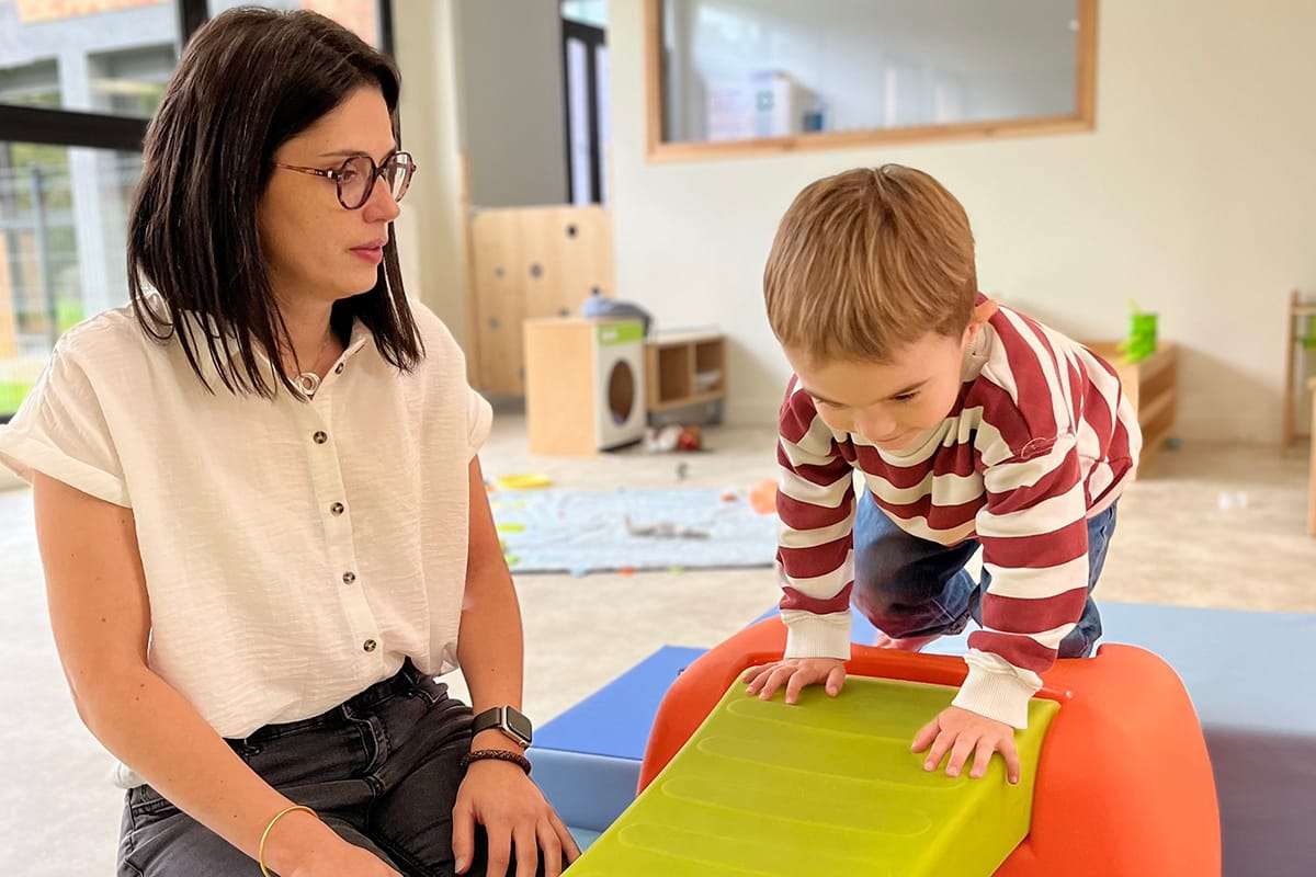 Un enfant grimpe sur un module de motricité coloré, accompagné d’une éducatrice qui l’encourage.