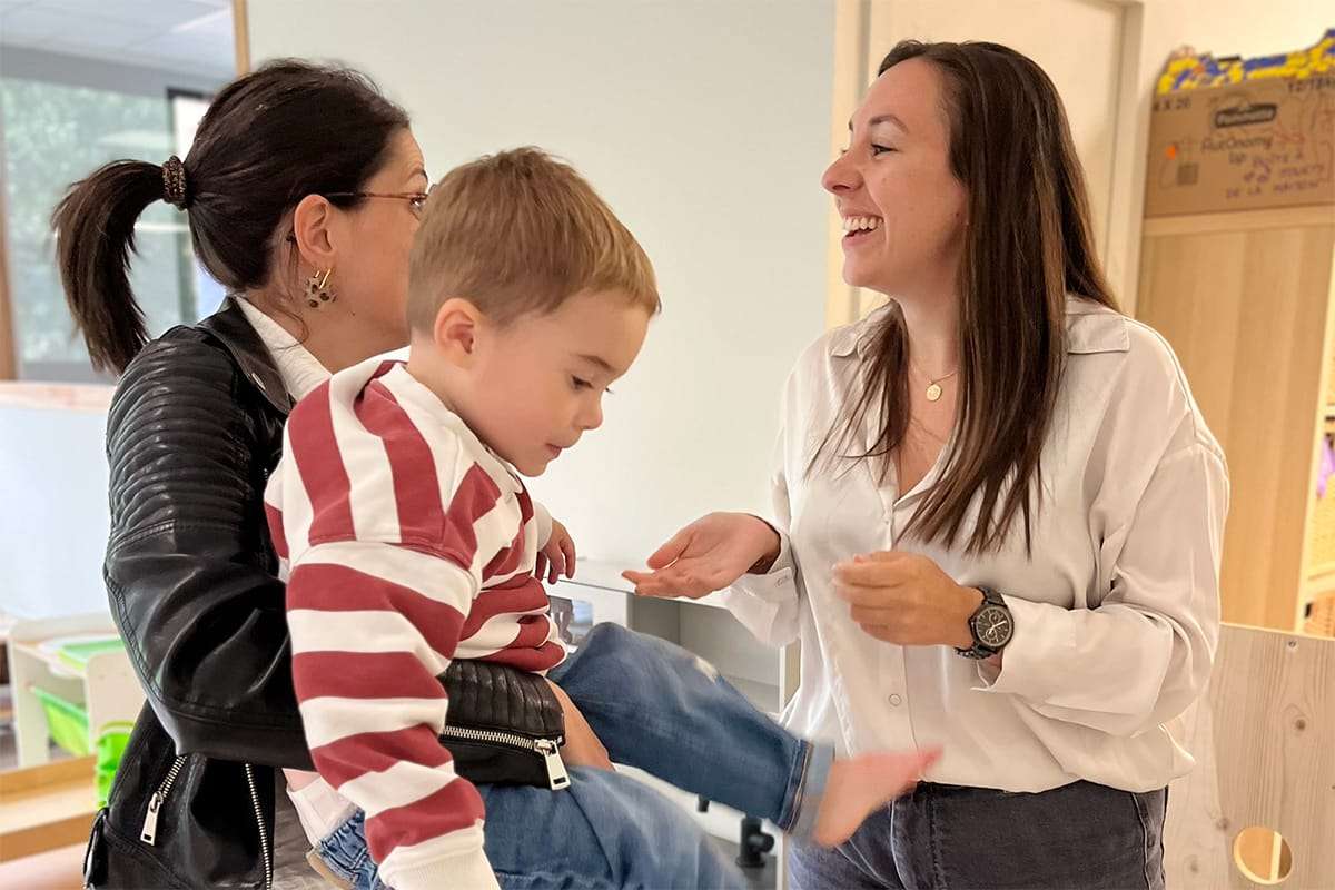 Une professionnelle de la crèche souriante, vêtue d'une chemise blanche, interagit avec une mère et son jeune enfant.