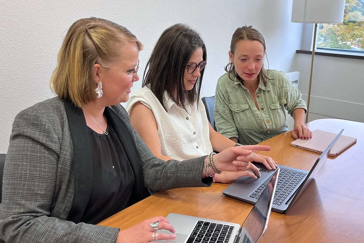 Plan sur trois femmes, membres de l'équipe de la crèche, assises autour d'une table en bois et travaillant ensemble sur des ordinateurs portables. La femme à gauche, vêtue d'une veste de costume grise, pointe du doigt l'écran d'un ordinateur. Les deux autres femmes regardent l'écran, engagées dans une discussion.