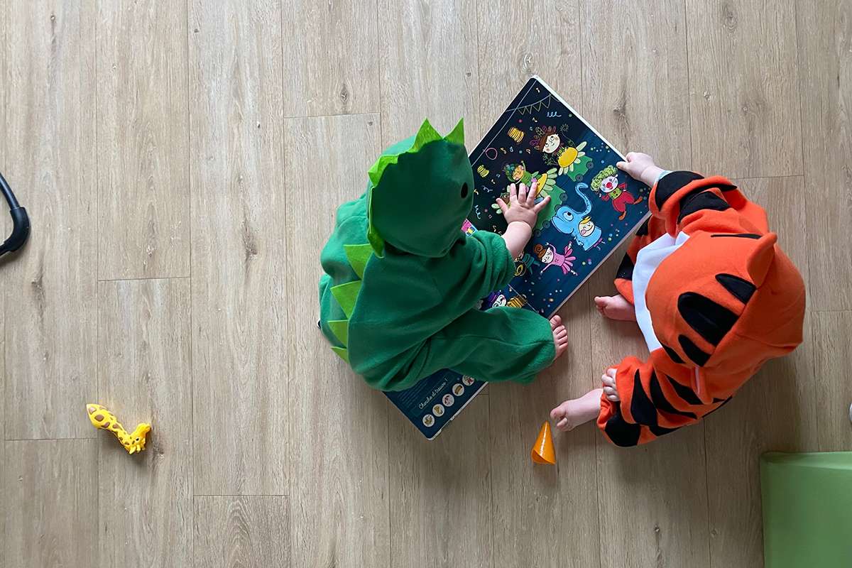 Vue de dessus de deux jeunes enfants vêtus de costumes d'animaux, assis sur un sol en bois clair. L'enfant de gauche porte un déguisement de dinosaure vert et l'enfant de droite un déguisement de tigre orange et noir. Ils sont penchés sur un livre ou un tableau d'activités illustré. Un jouet girafe est posé à côté d'eux sur le sol.