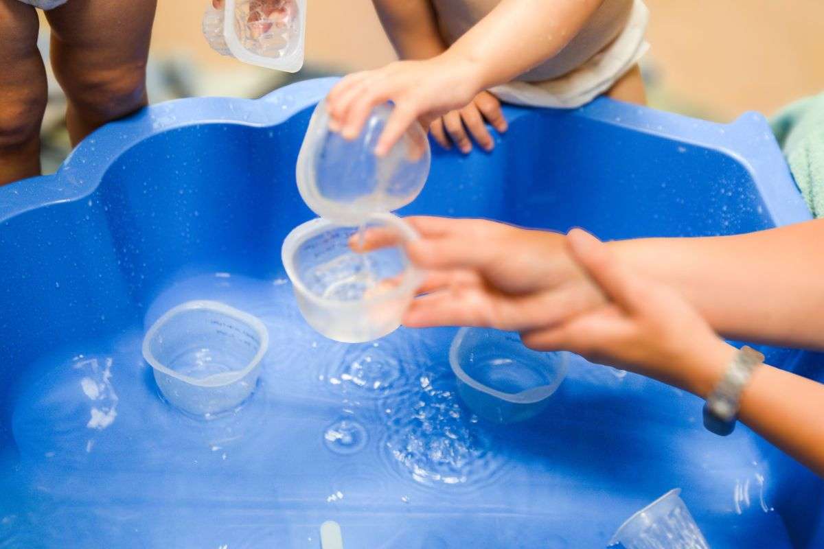 61 Des enfants en bas âge jouent avec de l’eau et des gobelets dans un bac bleu, sous la surveillance d’un adulte.